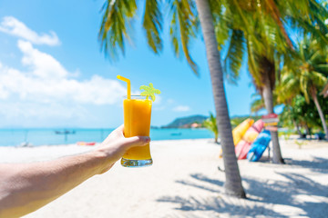 First-person view. Girl holds a glass cup of cold mango fresh on the background of a sandy tropical beach. White sand and palm trees. Fairytale vacation