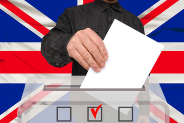 male voter drops a ballot in a transparent ballot box against the backdrop of the Great Britain national flag, concept of state elections, referendum