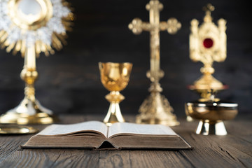 Catholic religion concept. Catholic symbols composition. The Cross, monstrance,  Holy Bible and golden chalice on wooden altar and gray background. 