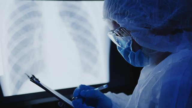 A woman in a medical protective uniform, goggles and a mask on her face examines an x-ray with pneumonia of the lungs on a monitor screen, writes information to a tablet