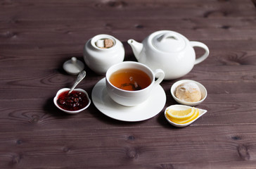  cup of herbal tea, teapot, sweet bun, sugar, raspberry jam, lemon on brown wooden background