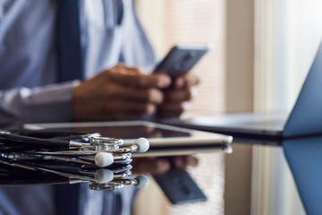 Male doctor in blue shirt with tie, hand holding and using mobile smart phone with laptop computer, medical stethoscope and digital tablet on the desk. Medical online networking, telehealth concept.