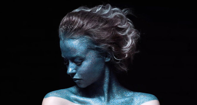 a girl with a blue face in profile in sparkles close-up against a black wall looks forward