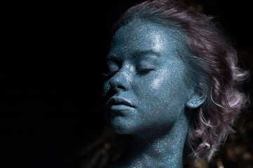 girl with a blue face in sparkles close-up against a black wall 