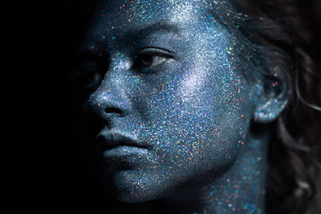 a girl with a blue face in profile in sparkles close-up against a black wall looks forward