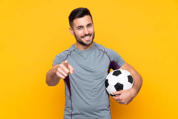 Man over isolated yellow background with soccer ball and pointing to the front