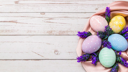 easter background with hand painted pastel colors eggs and fresh lavender flowers on pink silk fabric over light colored wooden table
