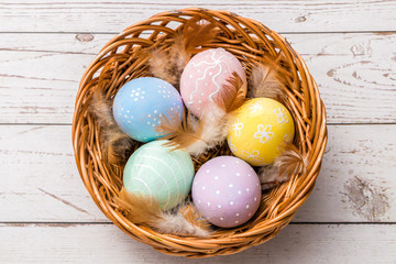 top view of easter eggs hand painted in pastel colors and tender feathers in a wicker basket on wooden table