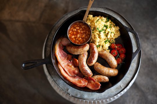 A full English breakfast in a frying pan with sausages, bacon, scrambled eggs, beans and tomatoes