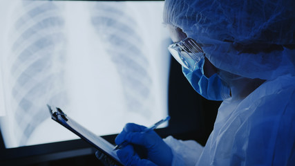 A woman in a medical protective uniform, goggles and a mask on her face examines an x-ray with pneumonia of the lungs on a monitor screen, writes information to a tablet