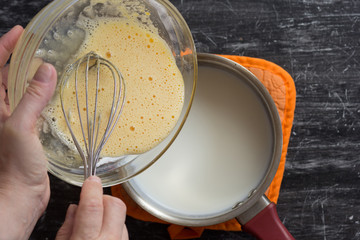 Top view of woman hands putting mix of yolks, starch, sugar and milk in milk for making custard cream on the black background