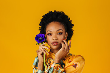 Portrait of an elegant  young woman holding a blue flower with orange finger nails, isolated on yellow background - Powered by Adobe
