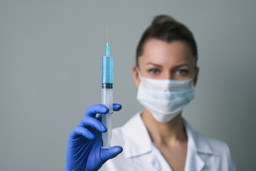 A female medical worker in a white robe and mask holds a syringe.