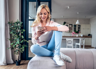 Attractive young woman spending time at home. Sitting on sofa with smart phone.