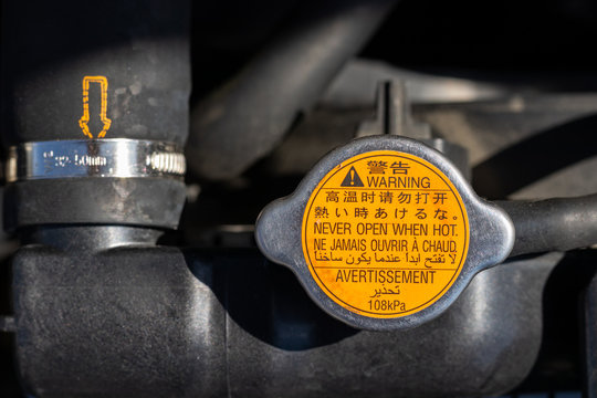 Orange radiator cap with warning labels in different languages: never open when hot, and blurred car radiator hose with yellow dotted arrow under the open hood of a car. Closeup view