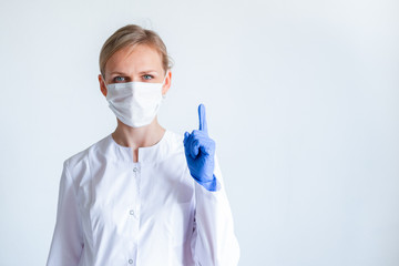 Attention. Blonde female doctor surgeon in white uniform, protective blue gloves and medical mask showing finger up over grey