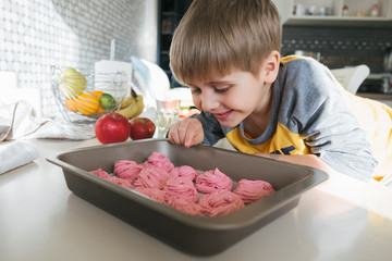 Boy 5 years old cheerful, smiling, wants to eat pink marshmallows. Home-cooked sweets, delicious food. Sitting in a sunny modern kitchen. Good mood.