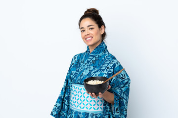 Woman wearing kimono over isolated white background smiling a lot while holding a bowl of noodles with chopsticks