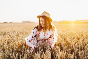 Naklejka na ściany i meble Young girl with long curly hairand freckles face, in hat, in light white dress with floral print, standing in wheat field, posing for camera, in background sundown.