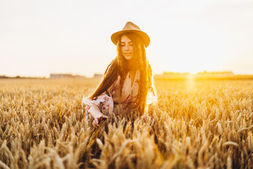 Young girl with long curly hairand freckles face, in hat, in light white dress with floral print, standing in wheat field, posing for camera, in background sundown.