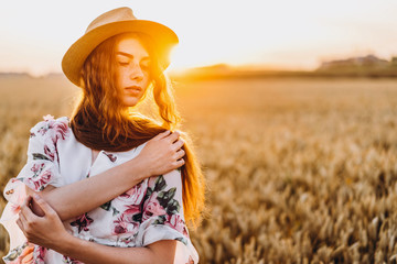 Portrait of a beautiful young woman with curly hair and freckles face. Woman in dress and hat posing in wheat field at sunset and looking at camera