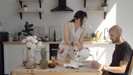 Beautiful girl or woman pours tea to her beloved husband in the modern kitchen. Quarantined family life due to coronavirus. Stay at home in comfort with your family.