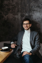 Young, successful businessman wearing glasses, gray jacket, white turtleneck on dark gray background