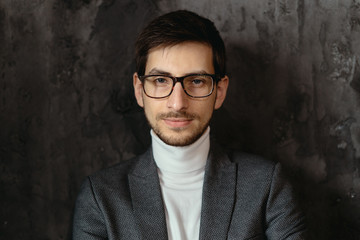 Young, successful businessman wearing glasses, gray jacket, white turtleneck on dark gray background