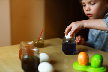 todder girl sitting at the table and dipping an egg in food paint making easter decoration