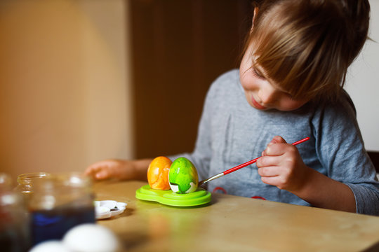 todder girl sitting at the table and painting easter eggs, holding brush in her hand