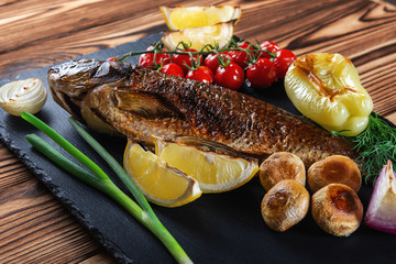 Grilled fish with vegetables, mushrooms and herbs on a black slate plate. Close up.