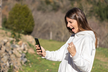 Beautiful euphoric brunette woman reads news on the smartphone in a park on a sunny day. She is...