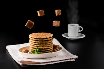 Several stacked stroopwafels or dutch waffles made from caramel syrup accompanied by a cup of steaming coffee and toffee candies
