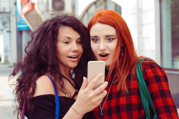 Closeup portrait two surprised girls looking at phone discussing latest gossip news Young shocked funny women friends reading sharing social media news on mobile cellphone pad computer in city outdoor