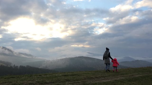 Happiness mother and daughter with backpack are running around the field on the grass mountains under the suns on clouds day. Camping on nature. 4k video. Carpathian autumn Mountains. Ukraine, Europe.