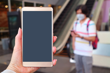Closeup blank mobile phone screen in woman's hand with blurry man wearing face mask to prevent infection in the backdrop