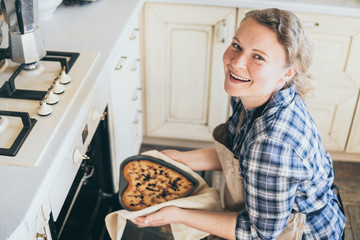 Young blonde woman taking heart shaped berry pie out of the oven. Cooking at home and smiling while...
