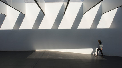 Una mujer pasando por un túnel con techo abierto que proyecta luces y sombras lineales
