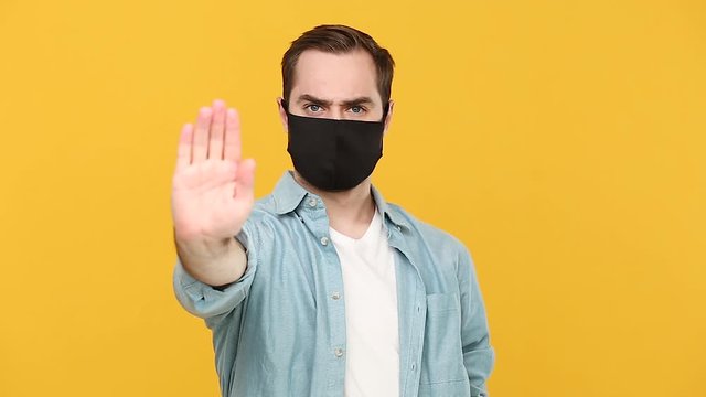 Young man in sterile black face mask denim white shirt posing isolated on yellow background studio. Epidemic pandemic coronavirus 2019-ncov sars covid-19 flu virus concept Sad guy showing stop gesture