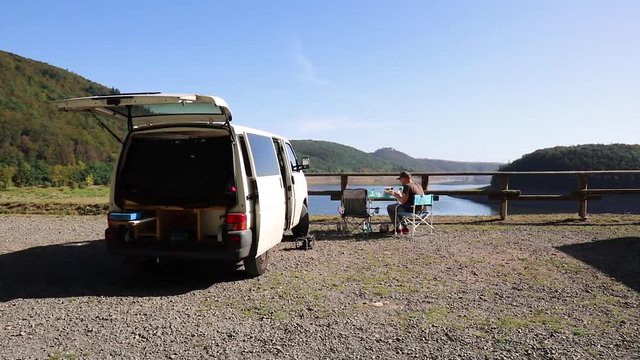Campervan at a lake surrounded by mountains / young man preparing a meal
