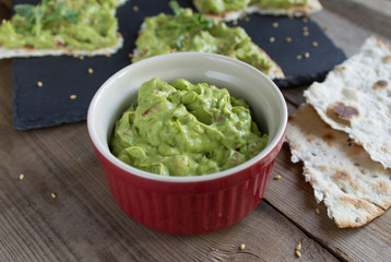 guacamole sauce in the red ramekin on a wooden surface with toasted tortillas on a black slate plate with sesame seeds