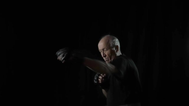 Elderly man in black t-shirt and gloves doing karate exercises on black background
