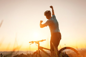 Man biker man meets a sunset in top of hill. He rising a one arm greeting on another successful day ending. Active sport people concept image