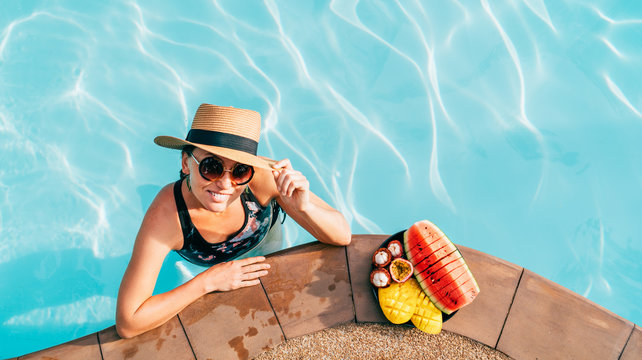 Soulful smiling woman in straw hat in sunglasses swimming in pool and enjoying fresh tropical fruits