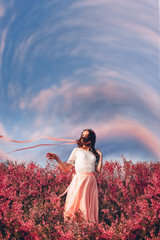 a beautiful girl in a dress in a field of pink flowers with a ribbon in her hair fluttering in the...