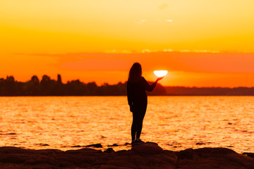 Girl standing on rock holding sun at hand. Nature and beauty concept. Orange sundown. Girl...