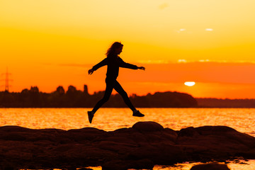 Young slim woman jumping on rock on sunset background. Nature and beauty concept. Orange sundown. Girl silhuette at sunset.