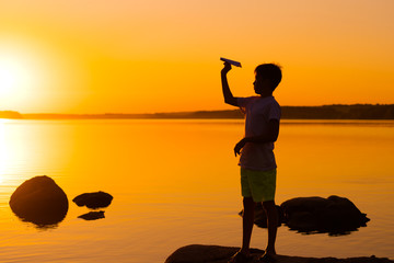 Teen boy looks at paper airplane in hand at orange sunset. A silhouette of a child with origami plane against water. Summer vocation.
