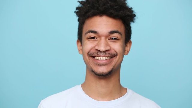 Unshaven african american young guy 20s in white t-shirt curly hair posing isolated on blue background studio portrait. People sincere emotions lifestyle concept. Looking at camera with charming smile