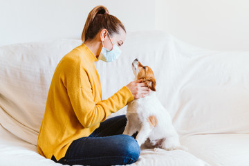 young woman hugging her cute small dog at home, sitting on the couch, wearing protective mask. Stay home concept during coronavirus covid-2019